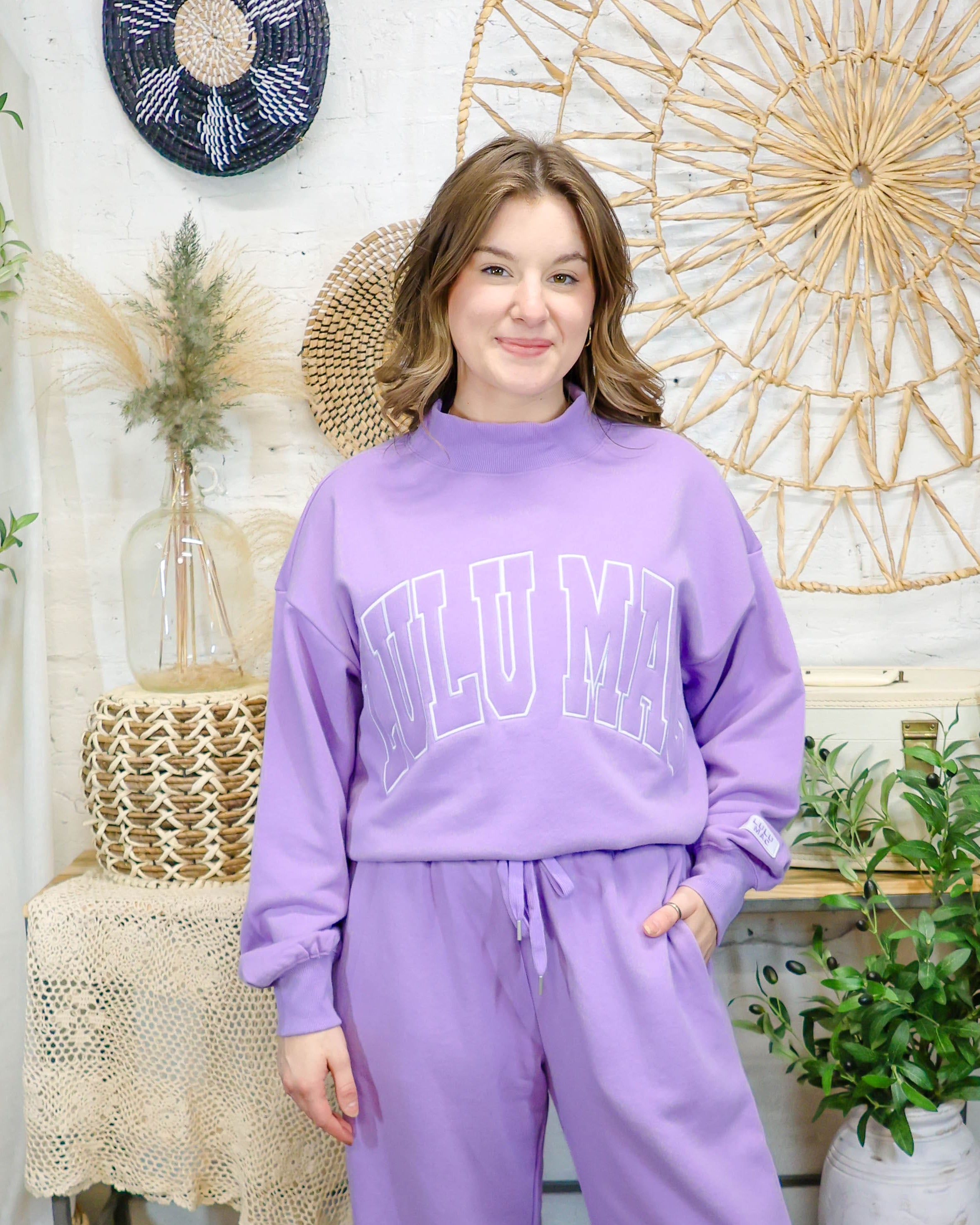 A woman modeling a matching lavender purple lounge set consisting of a sweatshirt with 'LULU MAC' text and wide-leg sweatpants, standing in a decorated boho-style room.